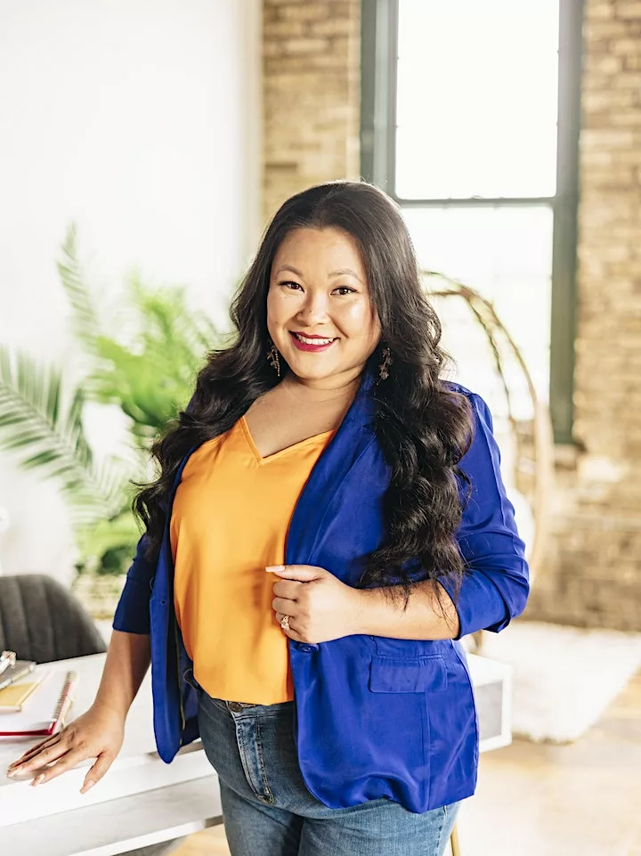 A professional portrait of Mollie Lo, a confident woman standing in a bright office space. She is smiling warmly at the camera, wearing a blue blazer over an orange top, paired with casual denim jeans. Her long black hair is styled in waves. Mollie rests one hand on a white desk and holds her other hand at her waist. Natural light streams in from a large window to her left, and there's a hint of greenery from an indoor plant in the background, contributing to a lively and welcoming atmosphere.