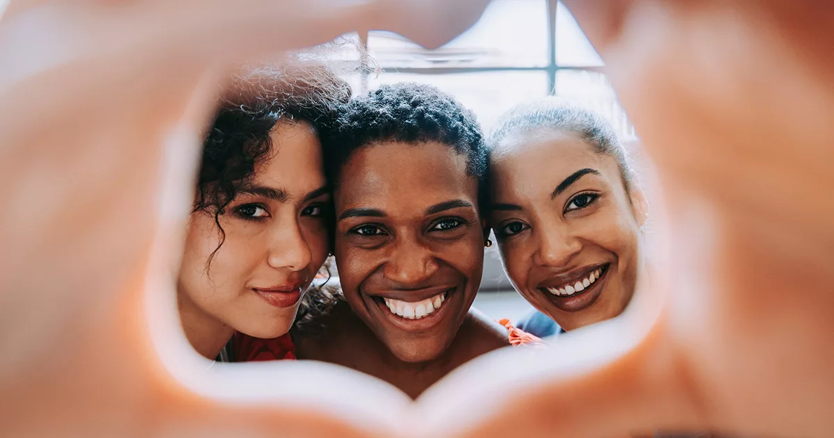 Three smiling friends of mixed ethnic backgrounds are seen up close, forming a heart shape with their hands, which frames their faces. They share a moment of joy and closeness, conveying a sense of friendship and love.