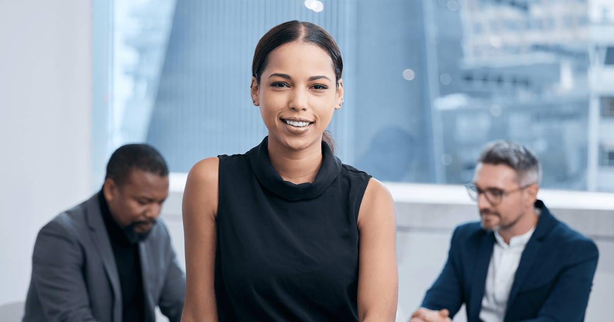 A confident businesswoman in a sleeveless black top smiles at the camera, ready to tackle the day's challenges, with two male colleagues in discussion at a table behind her in a modern office environment.