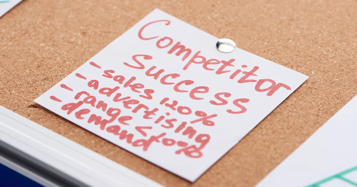 A close-up image of a red handwritten note pinned to a corkboard with the words 'Competitor Success' at the top. Below, a list includes '-sales 100%', '-advertising', '-range of products', and '-demand', suggesting a strategic analysis or performance metrics in a business context.