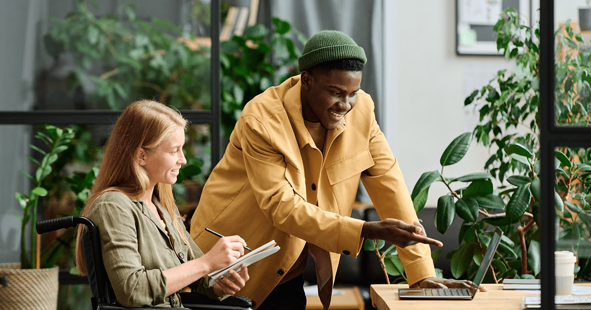 A young, confident businessman in a mustard jacket and green beanie points at a laptop screen, engaging with a colleague who is attentively taking notes in a wheelchair. They are in a bright, plant-filled office space, collaborating in an inclusive work environment.